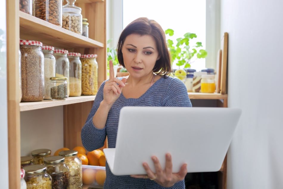 Woman in kitchen pantry with stored products, holding laptop