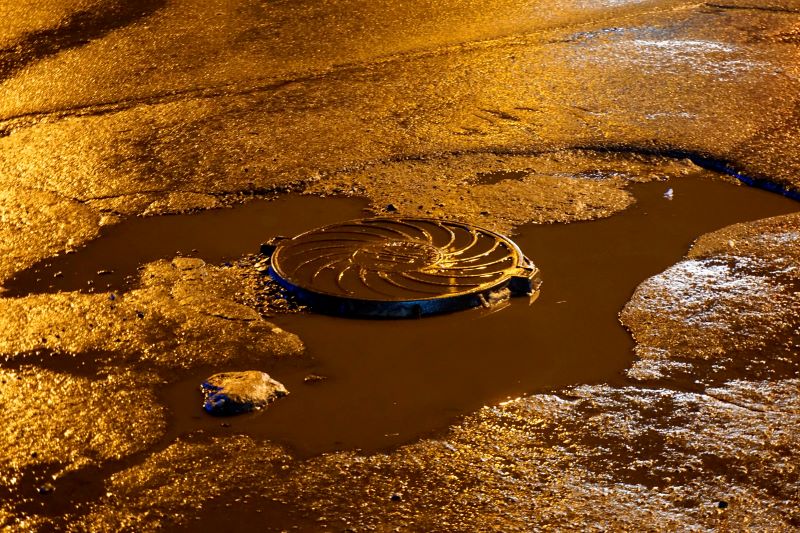Sewage hatch in a dirty puddle on the asphalt. Night yellow brown light