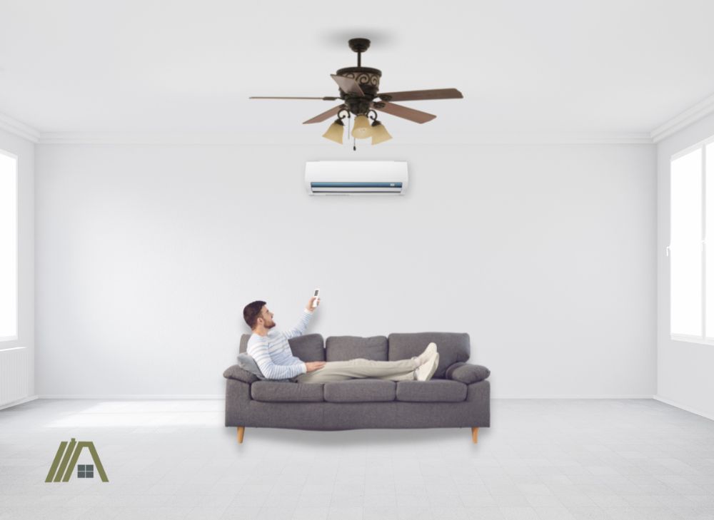 Man lying on a sofa holding a remote for the air-conditioning unit in a white room with ceiling fan