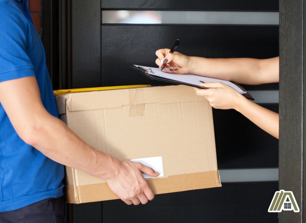 Man in blue shirt holding a package in front of a customer's door while the customer signs for the receiving list
